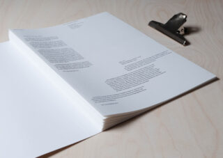 Ein Stapel bedruckter Papiere mit Text liegt offen auf einer hellen Holzoberfläche neben einer Klemmbrettklammer aus Metall.