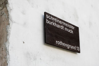 Auf einem dunklen Holzschild an einer weißen Wand steht in kleinen weißen Buchstaben „Schreinermeister Burkhardt Muck Rothengrund 13“.