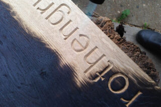 Eine Nahaufnahme eines Holzbretts mit dem eingeritzten Wort „lothengy“, aus einem bestimmten Winkel betrachtet, mit einem Teil eines Baumstamms und einem Bürgersteig im Hintergrund.