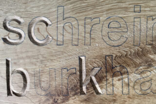 Nahaufnahme einer gravierten Holzoberfläche mit teilweise sichtbaren Wörtern „Schrei“ und „Burkha“ in umrissenen und geschnitzten Buchstaben.