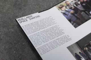 Eine Broschüre auf grauer Oberfläche zeigt den Text „Reallabor Space Sharing“ neben kleinen, unscharfen Fotos von Menschen in ungezwungener Atmosphäre.