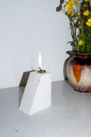 Eine kleine grüne Kerze brennt auf einem strukturierten weißen geometrischen Block, neben einer Keramikvase mit gelben Blumen auf einer hellen Oberfläche.