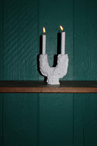Zwei brennende weiße Kerzen in einem strukturierten weißen Halter stehen auf einem Holzregal vor einer grün getäfelten Wand.