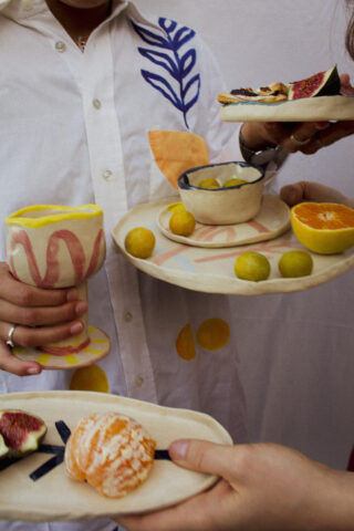 Zwei Personen halten Keramikteller und -tassen mit Feigen, Zitrusfrüchten und Snacks. Eine Person trägt ein weißes Hemd mit bunten abstrakten Mustern.