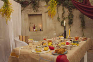 Ein rustikaler Tisch ist mit verschiedenen Gerichten, Obst, Kerzen und Getränken in einem Außenbereich mit Hängepflanzen und drapiertem Stoff gedeckt.
