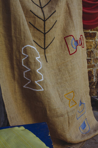 Ein hellbrauner Sackleinenstoff ist drapiert und mit abstrakten und geometrischen Stickereien aus schwarzem, weißem, rotem, blauem und gelbem Garn verziert. Im Vordergrund ist teilweise eine grüne Fläche sichtbar.