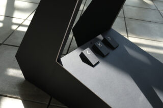 Eine Nahaufnahme von drei großen, schwarzen Tastaturtasten – Pfeil links, X und Pfeil rechts –, die auf einer abgewinkelten, mattschwarzen Oberfläche vor einem gefliesten Boden angebracht sind.