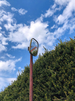 Vor hohen grünen Büschen steht ein konvexer Spiegel auf einer verrosteten Stange und reflektiert einen Teil eines Gebäudes mit rotem Dach unter einem blauen Himmel mit vereinzelten Wolken.
