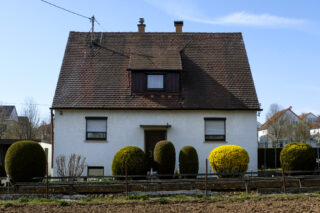 Ein zweistöckiges Haus mit einem steilen, braunen Ziegeldach, drei Fenstern und gestutzten Büschen im Vorgarten an einem klaren Tag.