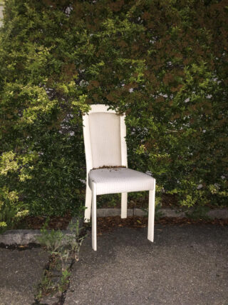 Ein weißer Stuhl steht vor einer grünen Hecke auf einer Betonfläche, auf der Sitzfläche liegen einige Blätter und Abfälle.