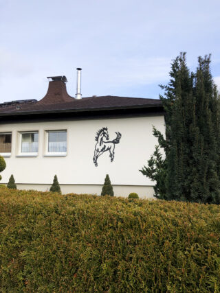 Ein weißes Haus mit einem schwarzen, sich aufbäumenden Pferd als Dekoration an der Außenwand, umgeben von grünen Büschen und hohen immergrünen Bäumen.