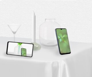 Zwei Smartphones zeigen ein Bild einer grünen Pflanze; ein Telefon steht aufrecht, das andere liegt horizontal. Sie liegen auf einer weißen Tischdecke neben Glaswaren und einer weißen Kerze.