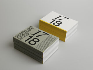 Zwei ordentlich gestapelte Taschenbücher mit fettgedruckten schwarzen Zahlen „17 18“ auf weißem Einband und gelbem Schnitt; ein Stapel hat graue Rücken mit der Aufschrift „GRUNDKLASSE PROF. GERWIN SCHMIDT“.