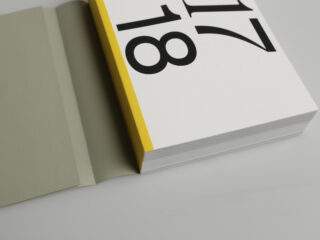 Ein dickes Buch mit gelbem Rücken und großen schwarzen Zahlen „17 18“ auf dem Einband liegt teilweise geöffnet auf einer hellgrauen Fläche.