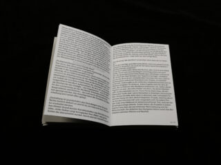 Auf einer schwarzen Fläche liegt ein aufgeschlagenes Buch mit dicht gedrucktem deutschen Text, fotografiert aus der Draufsicht.
