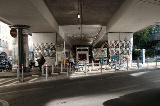 Fahrräder sind unter einer Überführung in der Nähe eines kleinen Gebäudes mit Plakaten und Graffiti an den Säulen und Wänden geparkt; Sonnenlicht wirft Schatten auf die Szene.