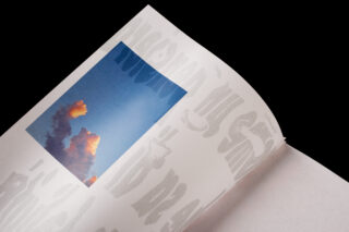 Ein offenes Buch zeigt auf einer Seite ein Foto von Wolken vor blauem Himmel, auf der angrenzenden Seite ist großer, stilisierter grauer Text abgedruckt.