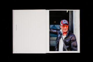 Offenes Buch mit einer leeren linken Seite und einem Foto auf der rechten Seite, das einen älteren Mann mit Mütze, Sonnenbrille, dunkler Pufferjacke und weißem Hemd zeigt, der mit einem erhobenen Arm dasteht.