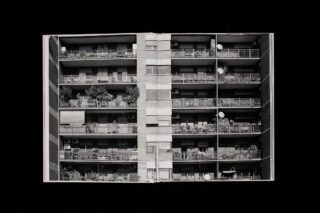 Schwarzweißfoto eines mehrstöckigen Wohnhauses mit Balkonen, Pflanzen und Satellitenschüsseln auf jeder Etage, von vorne betrachtet.