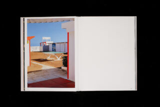 Offenes Buch mit einem Foto einer sonnenbeschienenen Außenszene mit weißen und roten Strukturen, blauem Himmel und sandigem Boden; die rechte Seite ist leer.