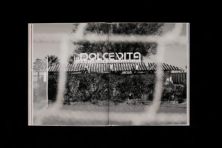 Schwarzweißfoto eines Gebäudes mit einem „DOLCEVITA“-Schild, durch einen Maschendrahtzaun gesehen; Bäume und Sträucher umgeben das Gebäude.