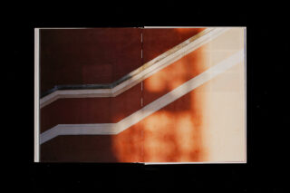 In einem aufgeschlagenen Buch ist die Fotografie eines diagonalen Treppengeländers zu sehen, das Schatten auf eine rotbraune und beige Wand wirft, wobei das Sonnenlicht auf der rechten Seite einen hellen Bereich erzeugt.