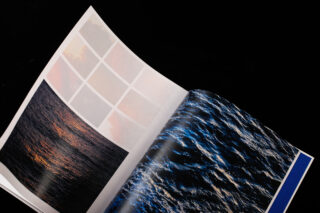 Eine aufgeschlagene Zeitschrift zeigt auf einer schwarzen Oberfläche ein Foto eines Sonnenuntergangs über dem Wasser und ein Foto von blauen Meereswellen, zusammen mit einem Raster aus verschwommenen Farbmustern.