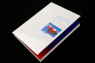 Ein geschlossenes Hardcover-Buch mit einem glänzend weißen Einband und einer kleinen Illustration eines Hamburgers und eines Getränks, fotografiert auf schwarzem Hintergrund.