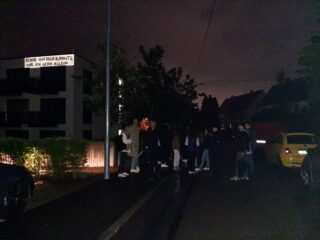 Eine Gruppe von Menschen steht nachts auf einer schwach beleuchteten Straße in der Nähe eines Wohnhauses, an dessen Wand ein beleuchtetes Schild mit deutscher Aufschrift hängt.