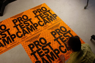 Eine Person kniet auf dem Boden und ordnet leuchtend orangefarbene Plakate an, auf denen in fetten schwarzen Buchstaben „PROTESTCAMP“ steht.