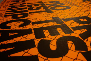 Große schwarze Buchstaben mit der Aufschrift „PROTEST“ und anderem Text sind auf einem orangefarbenen Hintergrund mit einem Maschendrahtzaunmuster gedruckt.