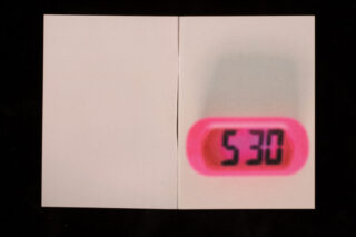 Ein kleiner digitaler Timer mit rosa Rand zeigt die Zahl 530 an und ist auf einer Fläche mit zwei weißen Blättern Papier platziert, alles auf schwarzem Hintergrund.