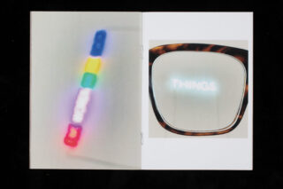 Auf einem aufgeschlagenen Heft ist links ein verschwommenes vertikales Neonschild mit farbigen Segmenten zu sehen, rechts spiegelt sich in einem Brillengestell das Wort „THINGS“.