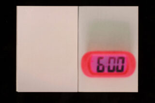 Eine kleine Digitaluhr mit rosa Rand zeigt neben einer leeren weißen Fläche die Uhrzeit 6:00 an.