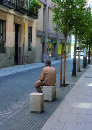 Ein älterer Mann in einer braunen Jacke sitzt allein auf einer Steinbank an einer ruhigen, leeren Stadtstraße, die von Bäumen und Gebäuden gesäumt ist.