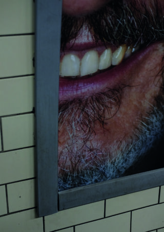 Nahaufnahme eines Posters, das die untere Hälfte des Gesichts eines lächelnden Mannes mit sichtbaren Zähnen und Bart zeigt, teilweise von einer Metallblende eingerahmt, an einer gefliesten Wand angebracht.