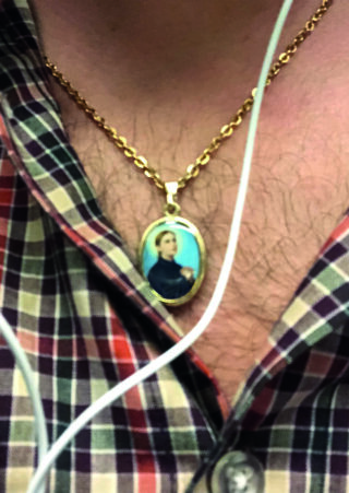 Über einem karierten Hemd wird eine goldene Halskette mit einem ovalen Anhänger getragen, der ein Frauenporträt zeigt; weiße Kopfhörerkabel sind sichtbar.