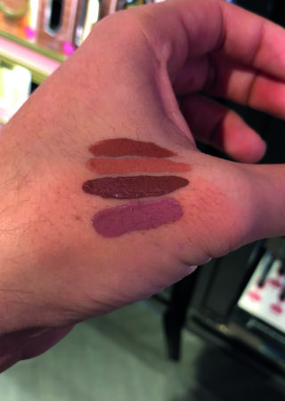 Eine Hand mit drei Farbmustern unterschiedlicher Lippenstift- oder Lipgloss-Töne auf der Rückseite, gezeigt unter Innenbeleuchtung.
