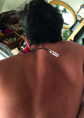 Eine Person mit kurzen dunklen Haaren und einer Halskette blickt von der Kamera weg und zeigt ihren nackten Rücken in einem Raum mit Regalen, Büchern und einem runden Spiegel.