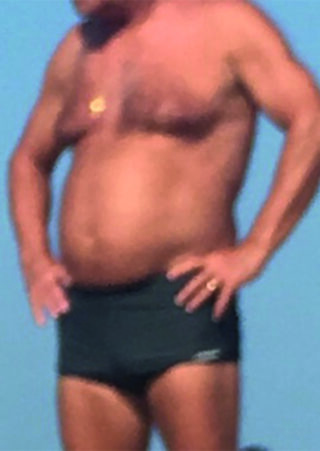 Ein Mann in einer schwarzen Badehose steht mit den Händen in den Hüften im Freien.