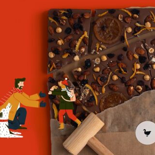 Schokoladenrinde mit Nüssen, Trockenfrüchten und Orangenscheiben auf braunem Papier, daneben ein Holzhammer und Comicfiguren auf rotem Hintergrund.