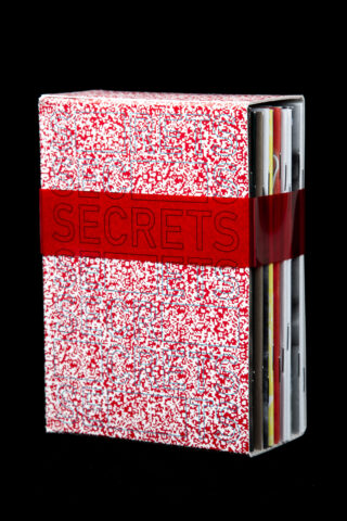 Eine Schachtel mit einem rot-weiß gesprenkelten Muster, die durch ein transparentes rotes Band mit der Aufschrift „SECRETS“ gesichert ist und mehrere Ordner oder Broschüren enthält.