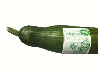 Eine Gurke mit einem Etikett mit der Aufschrift „vegane Wurst“, einer Zeichentrickfigur und dem Text „JETZT NEU!!“ auf schlichtem Hintergrund.