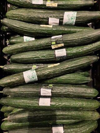 Ein Stapel langer, grüner Gurken mit der Aufschrift „Bio Gurke“ und einer Cartoon-Gurkenillustration auf einigen Etiketten.
