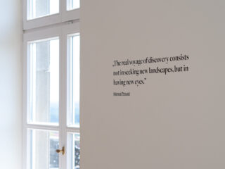 Auf einer weißen Wand ist ein Zitat von Marcel Proust über Entdeckungen zu sehen, neben einem Fenster, durch das Sonnenlicht fällt.