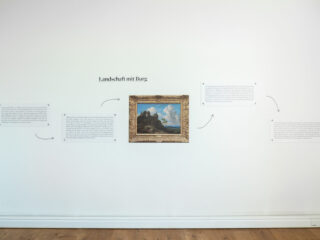 An einer weißen Wand hängt ein gerahmtes Landschaftsbild, darüber steht der Titel „Landschaft mit Burg“, umgeben von beschreibenden Texttafeln und Pfeilen, die auf das Kunstwerk hinweisen.