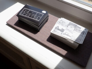 Zwei Stapel Postkarten liegen nebeneinander auf einer gewebten Matte neben einem Fenster. Ein Stapel weist ein geometrisches Muster auf, der andere zeigt Schwarzweiß-Fotoszenen.