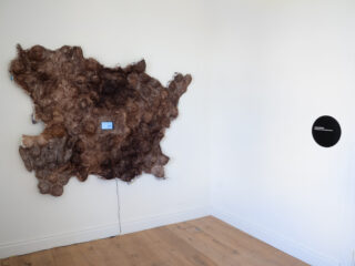 An einer weißen Wand ist ein Abschnitt aus strukturiertem braunem Material angebracht, an dem ein kleiner blauer Bildschirm und ein Kabel befestigt sind; an der angrenzenden Wand hängt eine schwarze runde Plakette.