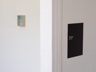 Eine weiße Wand mit einem schwarzen Schild mit der Aufschrift „Mini Les de rooij“ auf der rechten Seite und einer unscharfen kleinen Plakette an der linken Wand im Hintergrund.