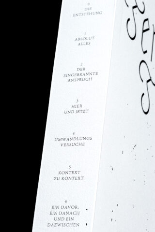 Nahaufnahme einer weißen Oberfläche mit einer nummerierten deutschen Textliste von 0 bis 6, wobei jedes Element eine kurze Phrase enthält und auf der rechten Seite teilweise schwarzer dekorativer Text vorhanden ist.